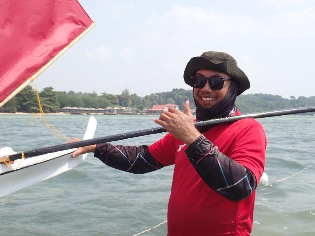 A racer in sunglasses and a headscarf holding a jong's white hull and red flag, giving a thumbs up on the water