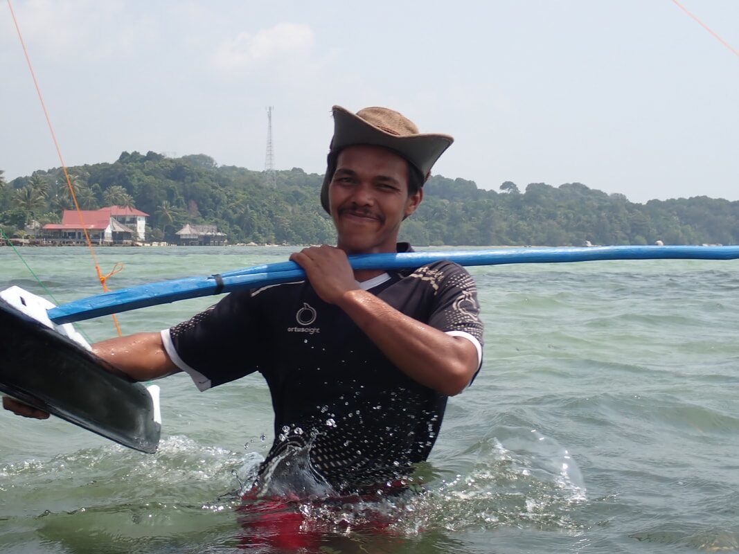 A player wading through the water carrying a jong's blue outrigger boom over his shoulder, resort buildings on the shore behind him