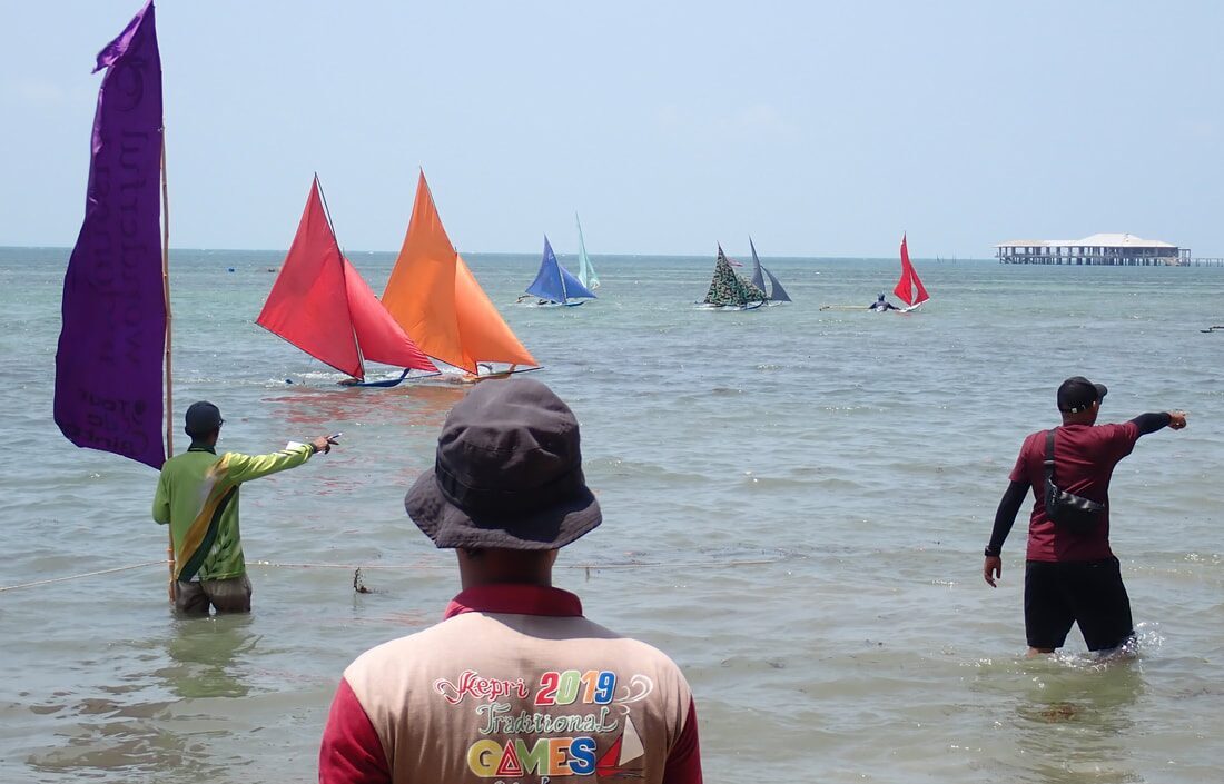 Race officials wading in the shallows watching red and orange-sailed jongs racing, a stilt building on the horizon