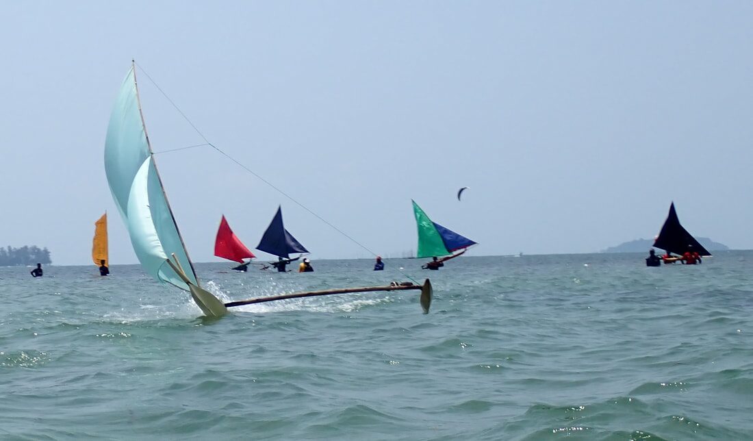 A pale teal-sailed jong throwing spray as it races, with orange, red, navy and green-sailed jongs strung out behind it