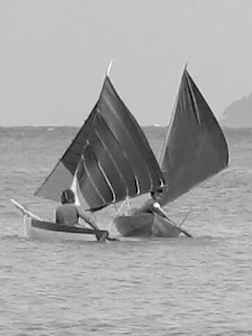Black-and-white photo of two small sampans with dark sails passing close together on the water, one rower aboard each