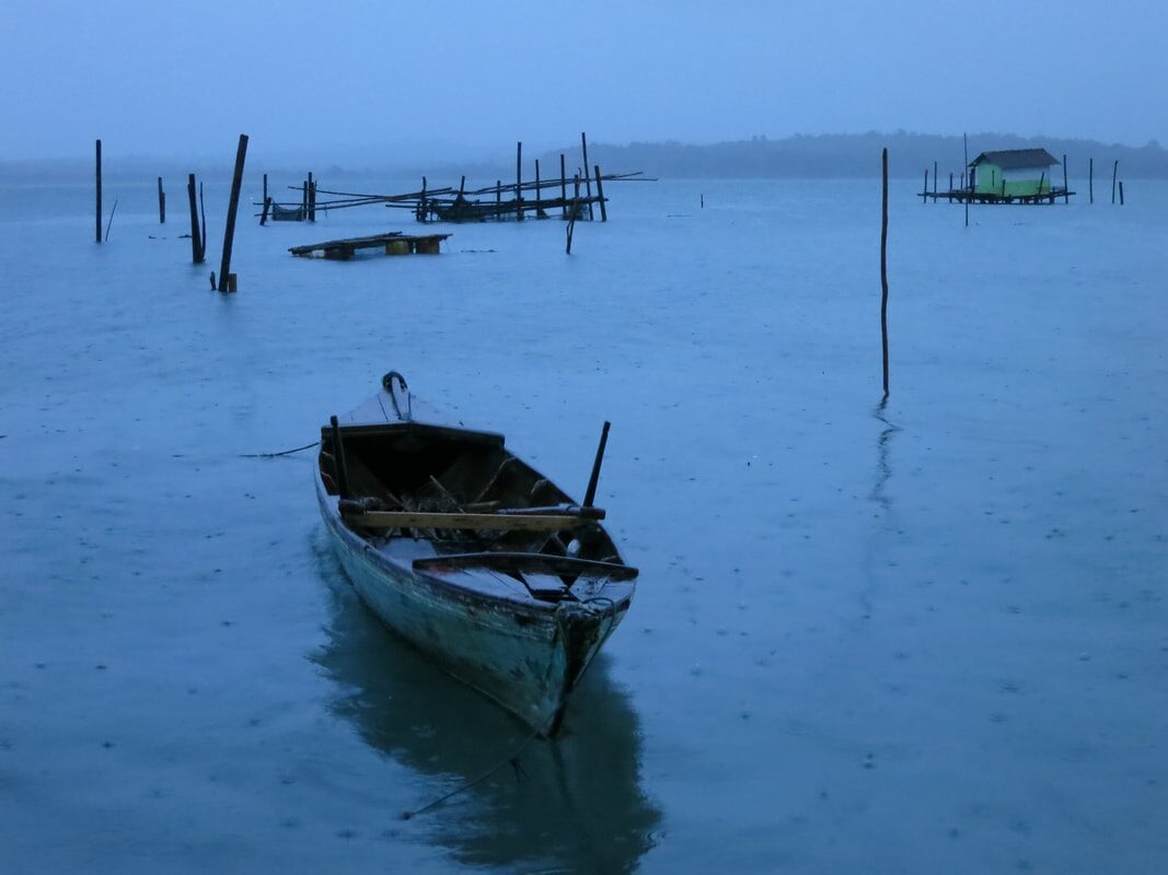 A lone wooden boat moored in the rain at dusk near Penyengat, a stilt hut and old fish-trap posts in the mist behind it