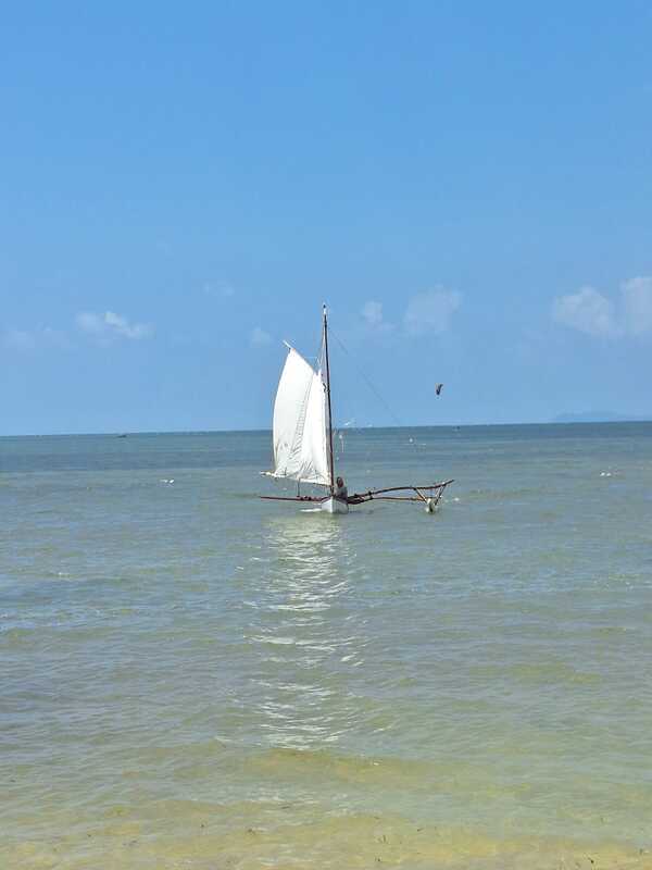 A small sampan layar with a single white sail resting near shore in shallow turquoise water
