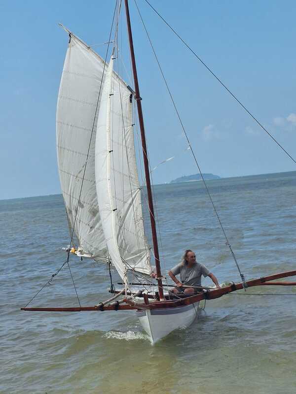 A man seated on the bow of a sampan layar under two full white sails, an island visible on the horizon