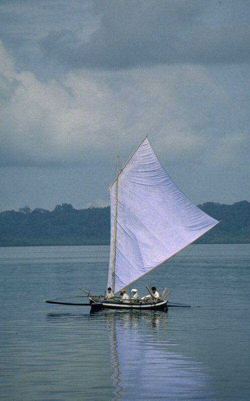 Archival photo of a traditional Sulawesi sailing boat with a tall white sail, three passengers aboard, sailing calm water