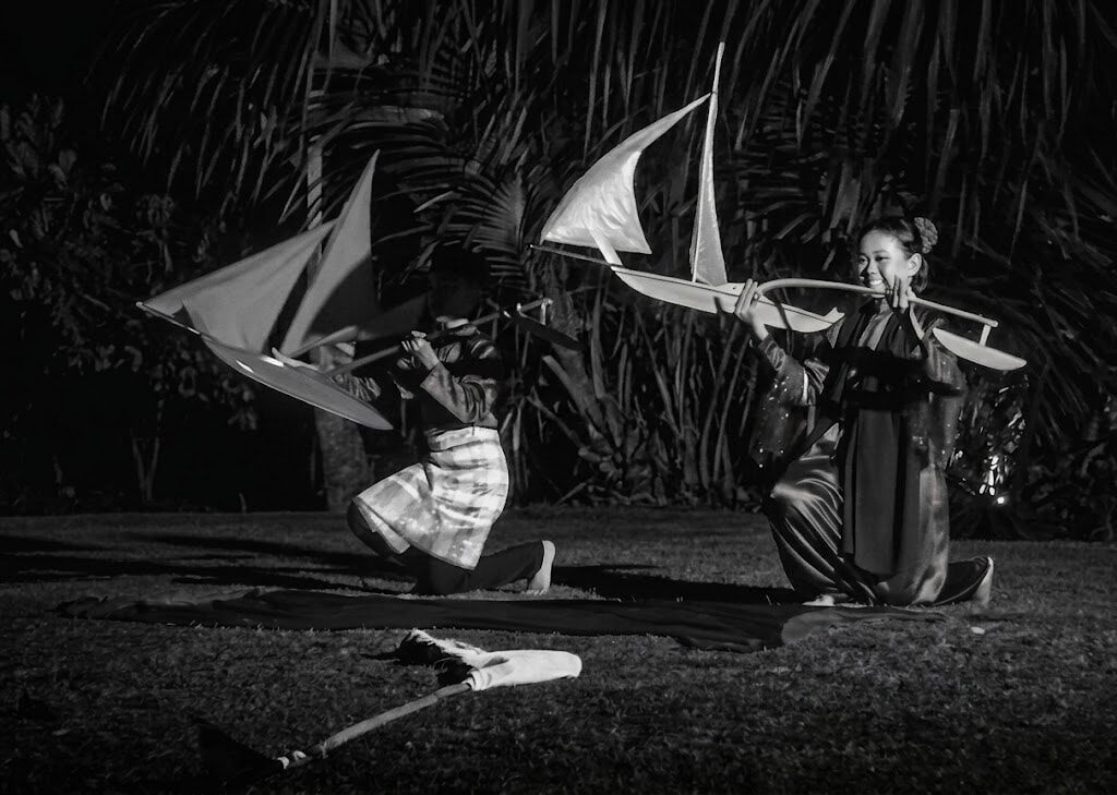 Black-and-white photo of two traditional dancers kneeling on a mat at night, each holding a model jong aloft against a backdrop of palm fronds