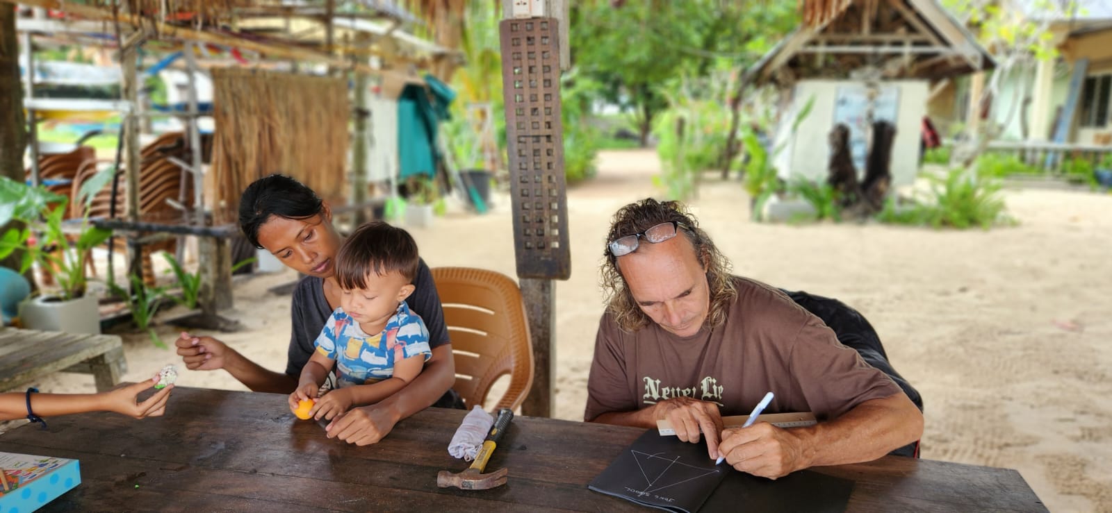Michel sketching jong rigging notes at a beach table, with a woman and a young child beside him