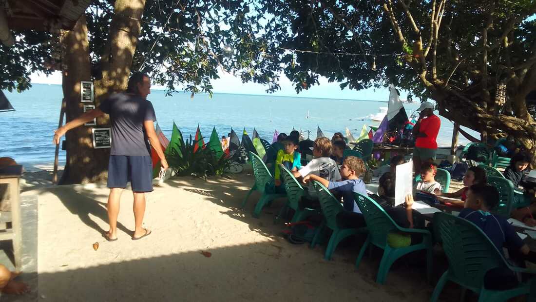 Michel talking with visiting schoolkids seated under a beachside tree at Planet Samudra, model jongs lined up along the shore behind them
