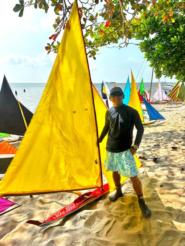 A player in a dark shirt and cap standing beside a jong with a bright yellow 'Nikoi Island' sail, other colorful sails on the beach behind him